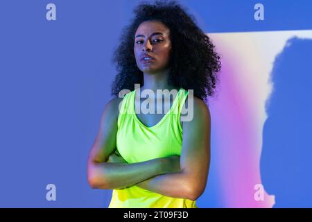Portrait jeune femme afro-américaine mince en vêtements d'activité debout avec les bras croisés sur fond violet regardant la caméra Banque D'Images