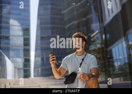 Touriste masculin avec bouteille d'eau se reposant après la promenade dans la ville moderne et faire la photo sur le téléphone Banque D'Images