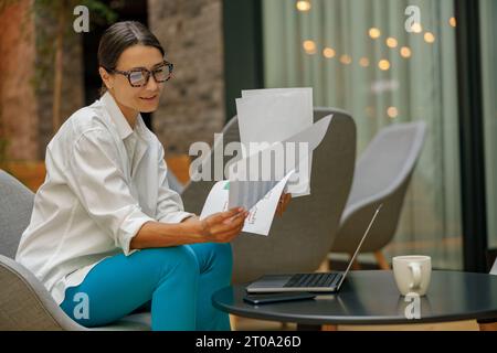 Jolie femme entrepreneure travaillant avec des documents et utilisant un ordinateur portable tout en étant assis dans le coworking Banque D'Images