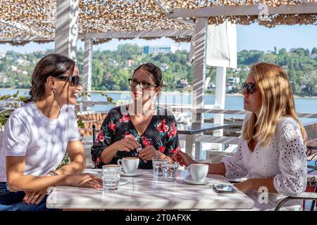 Trois amies joyeuses s'assoient à une table pour faire une pause café, bavarder et rire sur la terrasse d'un restaurant au bord de la rivière. Banque D'Images