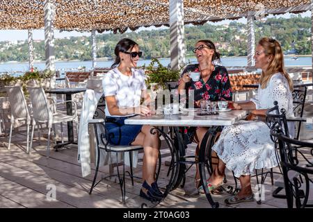 Trois amies joyeuses s'assoient à une table pour faire une pause café, bavarder et rire sur la terrasse d'un restaurant au bord de la rivière. Banque D'Images
