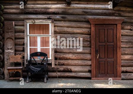 Fragment d'une maison en rondins avec une fenêtre rectangulaire dans un cadre blanc, une porte en bois et une chaise en cuir. Banque D'Images