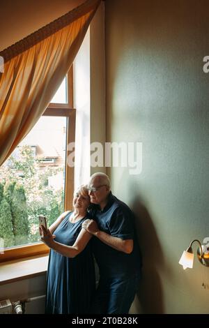 Un couple âgé heureux se tient près de la fenêtre, pose, prend un selfie au téléphone. Banque D'Images