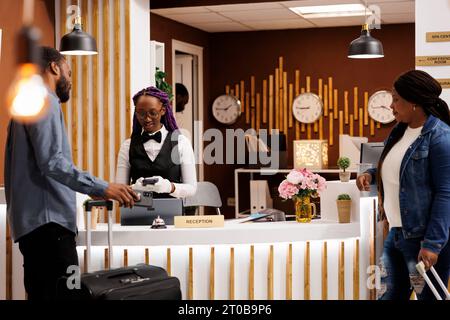 Jeune couple afro-américain debout à la réception payant pour la chambre d'hôtel lors du départ. Touristes utilisant le téléphone portable pour payer, en faisant le paiement nfc tout en s'inscrivant à la réception Banque D'Images