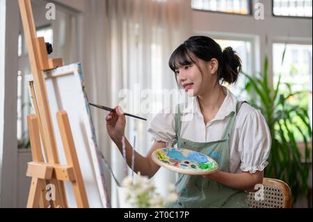 Une belle et heureuse jeune artiste asiatique aime peindre sur une toile dans son home studio tout en travaillant sur son projet artistique. passe-temps créatif Banque D'Images