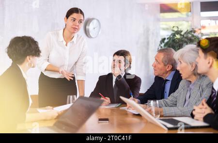 Intelligent jeune femme faisant rapport tandis que les employés de bureau d'âges différents utilisant des cahiers à la table dans le bureau Banque D'Images