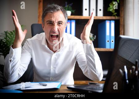 Homme d'affaires parlant au téléphone en colère et criant dans son bureau assis avec des documents sur la table dans le bureau avec des meubles en bois Banque D'Images