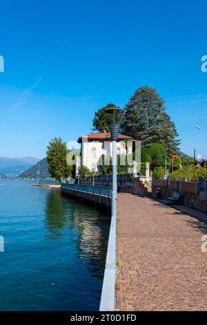 Passerelle avec lampes et balustrade et une maison sur le front de mer sur le lac de Lugano avec montagne et arbres dans une journée d'été ensoleillée à Porto Ceresio, Lombardie Banque D'Images