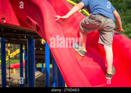 Jeune garçon grimpant le toboggan dans le terrain de jeu de banlieue. Banque D'Images