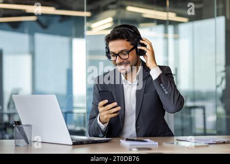 Jeune homme d'affaires joyeux se relaxant dans le bureau, homme dans les écouteurs écoute de la musique, chante et danse, utilise l'application sur le téléphone, travaille avec un ordinateur portable avec smartphone dans les mains. Banque D'Images