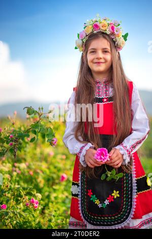 Fille bulgare avec la couronne de fleurs cueillant l'huile portant des roses damascena sur un champ de roses en fleurs au lever du soleil Banque D'Images