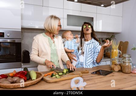 Grand-mère mère mère et petite-fille de bébé cuisinant ensemble dans la cuisine dans l'appartement tandis que maman faisant un drôle de visages pour l'enfant et la grand-mère coupant le végétab Banque D'Images