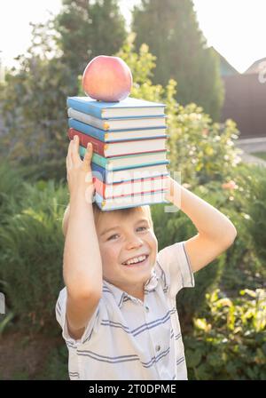 mignon et intelligent garçon tient une pile de livres sur sa tête et une pomme sur eux et rit joyeusement le premier jour de l'école. Retour à l'école, intérêt Banque D'Images