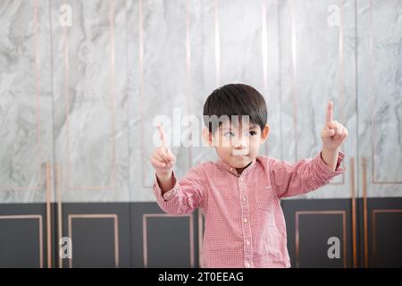 Un garçon asiatique se tient debout et sourit, levant les deux mains et pointant vers le haut. Banque D'Images