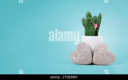 Pierre de message de rêve et de croire devant le cactus dans le vase sur fond bleu. Rochers en forme de cœur avec message de mot d'inspiration sculpté, osez à drea Banque D'Images