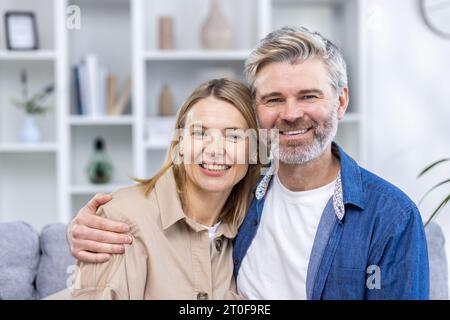 Portrait d'un couple mûr aux cheveux gris, homme de famille heureux et femme souriant et regardant la caméra, assis sur un canapé dans le salon à la maison. Banque D'Images