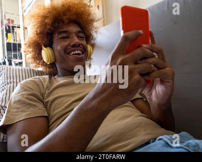 Homme afro-américain bavardant joyeusement avec smartphone allongé sur le canapé dans un appartement Banque D'Images
