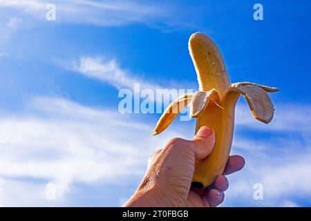 banane pelée à moitié mûre tenue par la main d'une femme sur fond de ciel bleu avec des nuages Banque D'Images