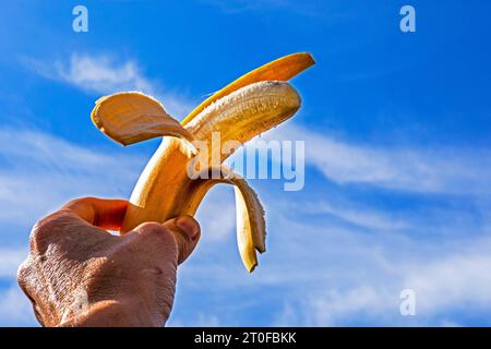 banane pelée mûre tenue par la main d'une femme sur le fond d'un ciel bleu avec des nuages Banque D'Images