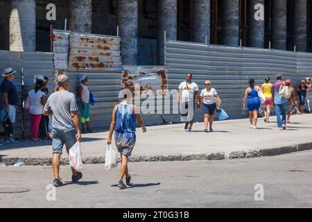 Des Cubains tenant des sacs à provisions en plastique marchent devant un vieux bâtiment en reconstruction ou en réparation. La structure est protégée par une clôture métallique. Banque D'Images