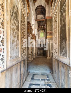 Couloir étroit avec des murs en marbre décorés dans le complexe Qalawun, un complexe islamique historique situé sur la rue Moez au Caire, en Égypte. Le couloir dispose d'un beau vitrail à une extrémité Banque D'Images