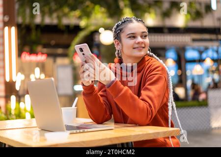 Fille alternative avec des tresses blanches en utilisant un ordinateur dans un centre commercial, souriant faire un message texte Banque D'Images