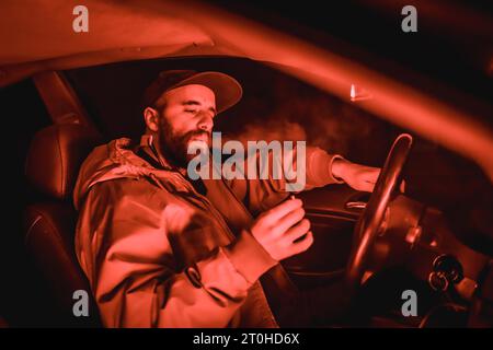 Rappeur homme dans une casquette dans une voiture fumant la nuit dans un garage éclairé avec une lumière rouge en hiver, voiture de sport Banque D'Images