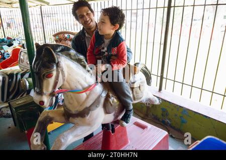 Vénézuélien Latino enfant mâle, heureux de monter sur le cheval prétendu avec son père à la foire de carrousel, concept de famille, espace de copie. Banque D'Images