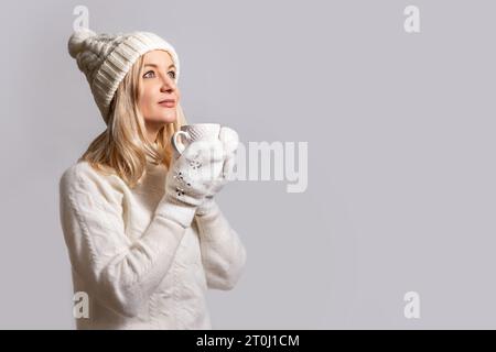 Femme en pull blanc en laine chaude, casquette avec bubo et mitaines tient la tasse, sourit et regarde vers le haut de rêve. Photo de studio sur fond clair. Banque D'Images