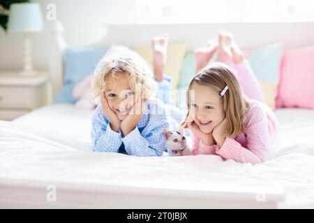 Les enfants jouent avec un chat dans un lit dans une chambre blanche. Enfant tenant un chaton blanc. Petite fille et garçon en pyjama avec un animal de compagnie mignon à la maison. Banque D'Images