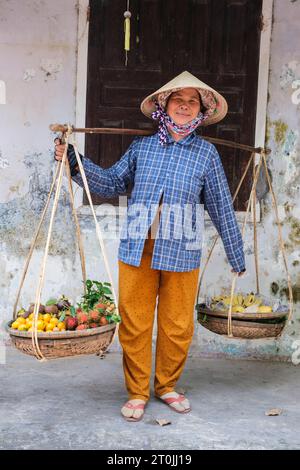 Hoi an, Vietnam. Femme portant des fruits tropicaux sur des poteaux d'épaule. Banque D'Images