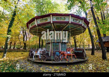 Vieux carrousel en bois peint pour enfants entouré de vieux grands arbres jaunes et verts dans le Musée du village dans le Parc Herastrau à Bucarest, Roumanie dans un Banque D'Images