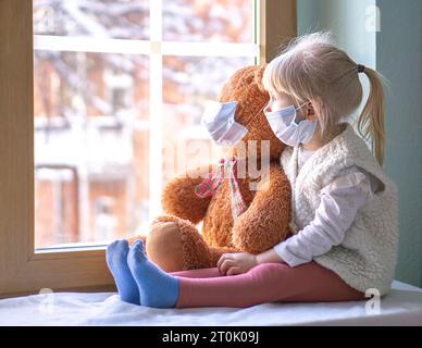 Petite fille et ours en peluche en masque assis devant la fenêtre et regardant dehors à la neige. Banque D'Images