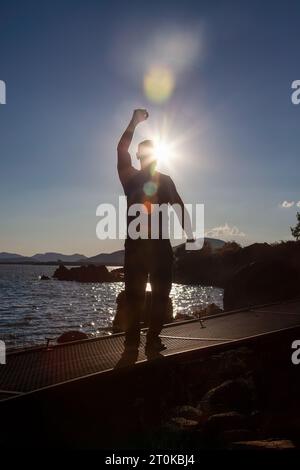 silhouette jante lumière au coucher du soleil rayonne l'homme avec la main gagnante levée sur un pont au lac Banque D'Images