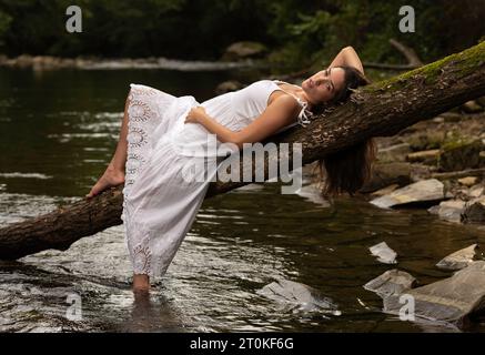 belle femme en robe blanche couchée sur une bûche sur la rive de la rivière Banque D'Images