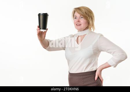 Femme blonde tenant tasse jetable en papier noir pour le café ou le thé dans la main tendue et regardant la tasse. femme de 49 ans vêtue d'un chemisier blanc Banque D'Images