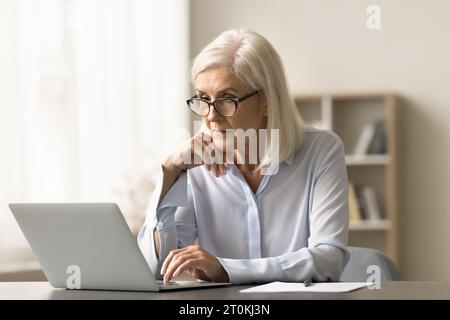 Femme d'affaires âgée concentrée dans des lunettes travaillant à l'ordinateur portable Banque D'Images