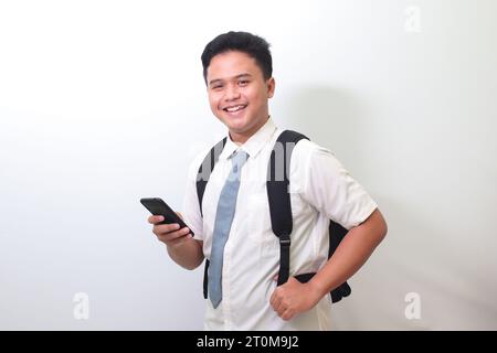 Étudiant de lycée indonésien portant un uniforme de chemise blanc avec cravate grise montrant une expression de visage choqué tout en tenant un téléphone portable. Isolé Banque D'Images