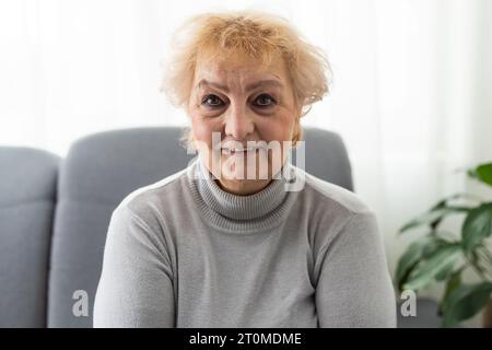 Heureuse dame âgée joyeuse regardant la caméra avec un sourire toothy, riant, assis sur la tête du canapé a pris le portrait. Femme assez âgée parlant sur la vidéo Banque D'Images