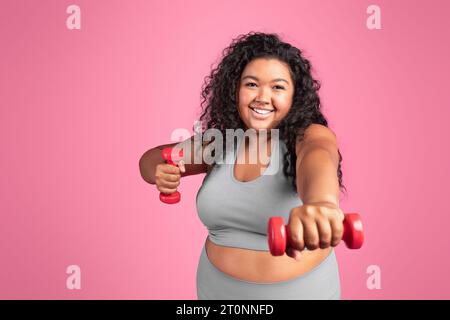 Joyeuse dame noire excitée Chubby en vêtements de sport faisant des exercices avec des haltères de main, debout sur fond de studio rose Banque D'Images