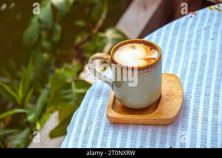 Une tasse chaude de cappuccino frais agrémentée d'un délicat latte art est posée sur un dessous de verre en bois au-dessus d'une nappe rayée. Banque D'Images