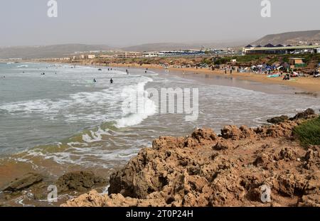 Surfeurs et amateurs de plage un jour de février à Tamraght près des hôtels en bord de mer de Taghazout Bay, NR Agadir, Maroc, photographiés depuis un promontoire rocheux. Banque D'Images