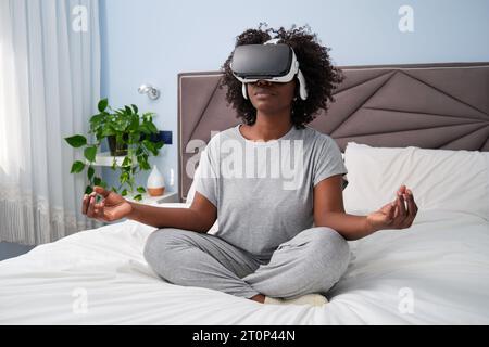 Jeune femme africaine dans le casque de réalité virtuelle méditant, appréciant la simulation et se relaxant dans la chambre à coucher. Banque D'Images