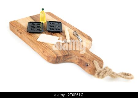 Set de cuisson de poupée avec moules à muffins, huile, fouet, planche à découper et rouleau à pâtisserie sur une plus grande planche en bois isolée sur blanc Banque D'Images