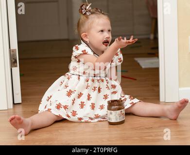 Une petite fille de 3 ans dans une belle robe blanche est assise sur le sol dans un intérieur de la maison et mange joyeusement du chocolat à tartiner dans un pot. Toute sa mout Banque D'Images