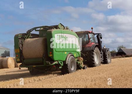 L'arrière d'une ramasseuse-presse McHale et d'un tracteur Red Valtra produisant des balles de paille d'orge sur un champ de chaumes à la fin de la récolte Banque D'Images