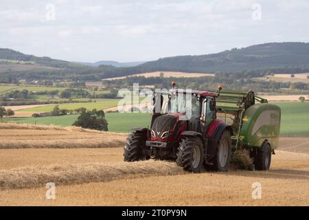 Tracteur Valtra rouge avec presse McHale travaillant dans un champ de chaumes avec vue sur la campagne pittoresque de l'Aberdeenshire Banque D'Images