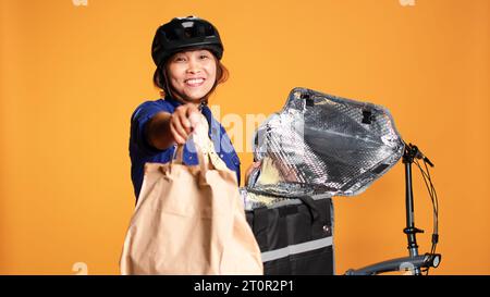 Photo POV du coursier arrivant à l'adresse du client, sortant le sac à lunch du sac à dos thermique. Joyeux cycliste asiatique livrant de la nourriture à emporter au client, isolé sur fond de studio Banque D'Images