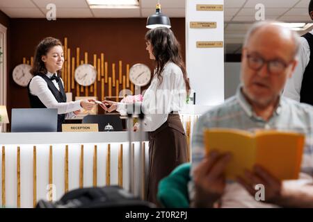 Femme caucasienne âgée arrive à la réception de l'hôtel et s'enregistre avec un document personnel. Au comptoir d'enregistrement, concierge féminin recevant un document d'identité de la voyageuse senior pour la réservation de chambre. Banque D'Images