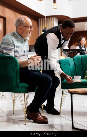 Dans le salon, un bon serveur servant une tasse de thé aux touristes masculins âgés assis sur un canapé avec tablette. Ancien homme à la retraite assis sur un canapé confortable et assisté par le personnel afro-américain dans le hall de l'hôtel. Banque D'Images